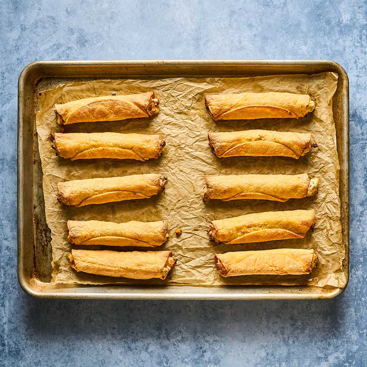 A baking sheet lined with parchment paper holds ten golden-brown baked taquitos arranged in two neat rows on a textured blue-gray surface.