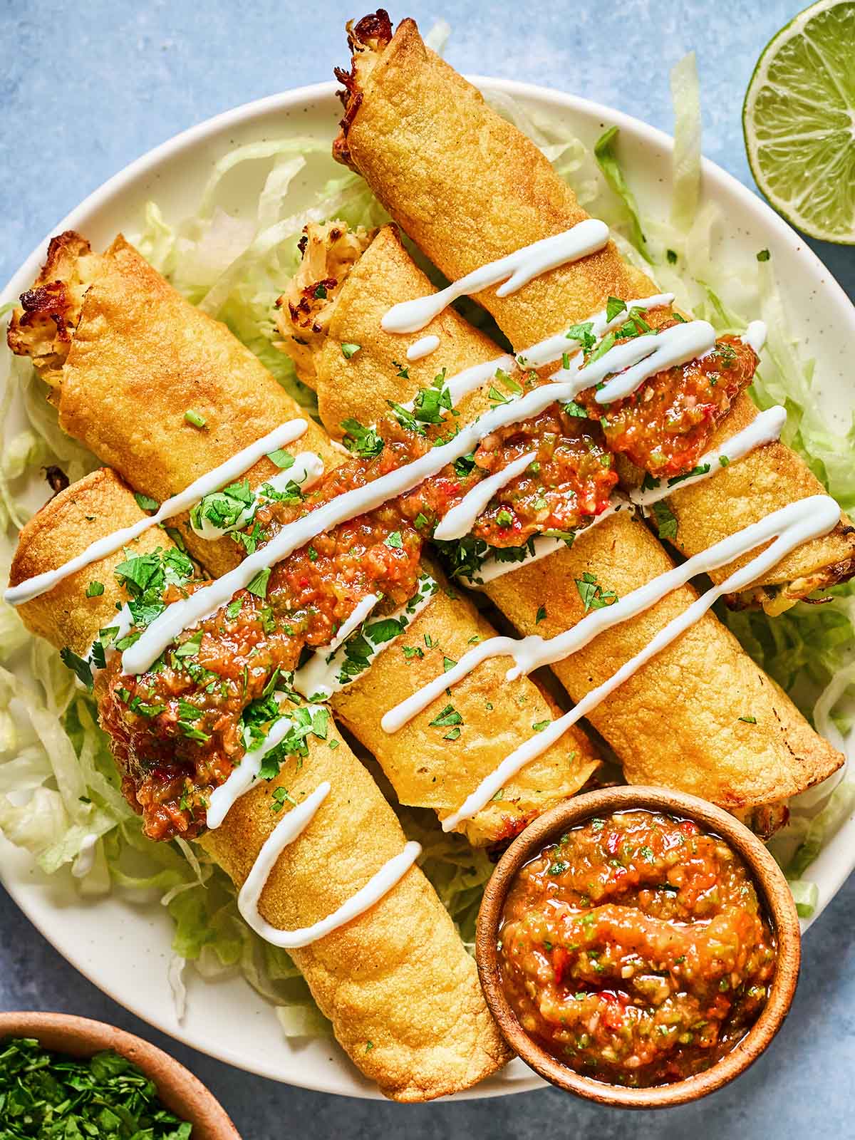 A plate of golden, crispy taquitos topped with salsa, sour cream, and chopped cilantro, served on a bed of shredded lettuce with a small bowl of salsa on the side.