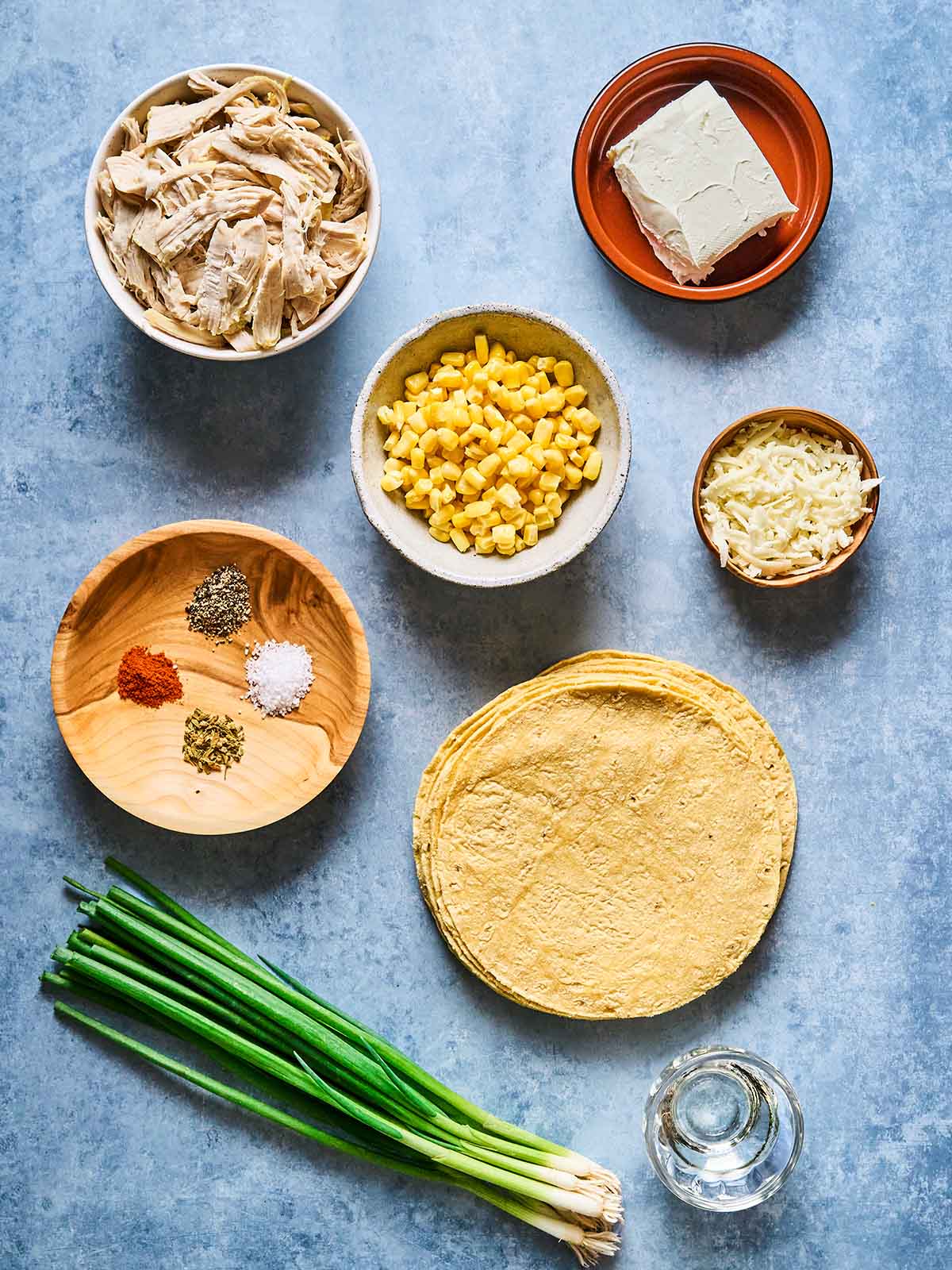 Assorted ingredients on a blue surface including shredded chicken, corn, cream cheese, shredded cheese, spices, corn tortillas, green onions, and a small glass of water.
