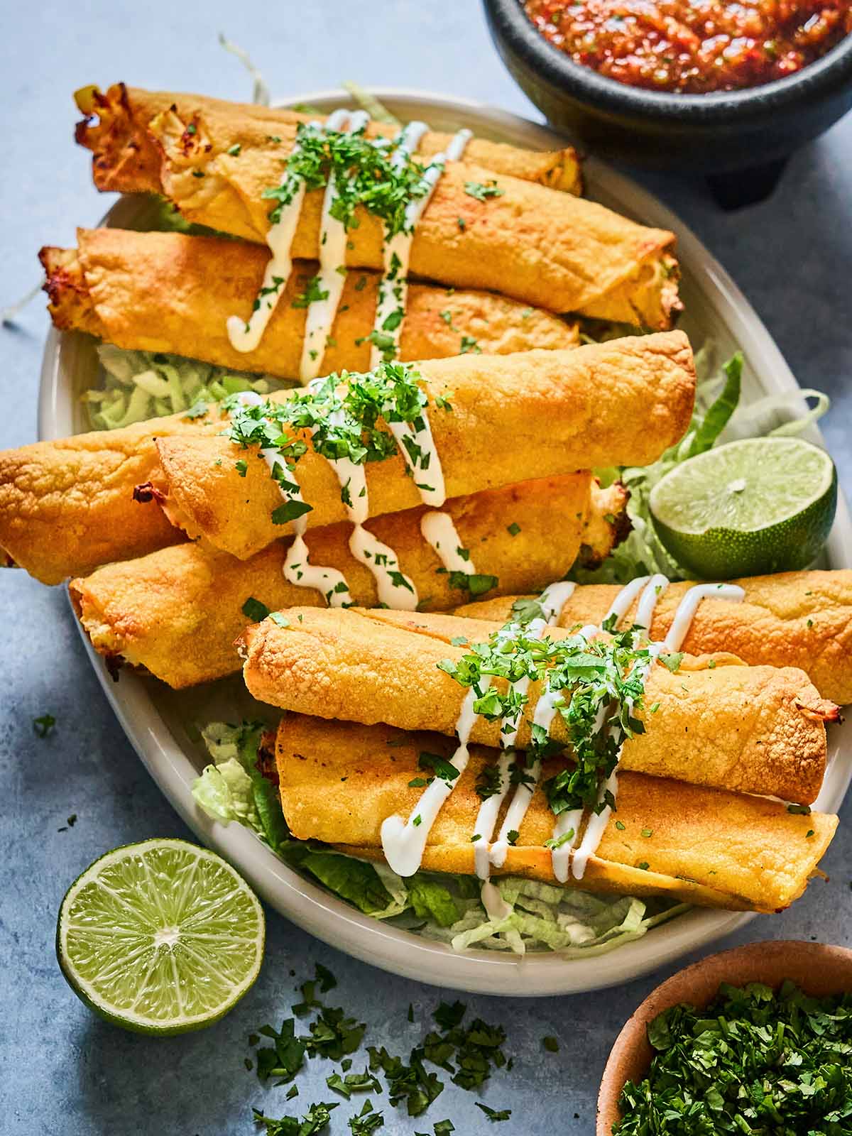 A plate of crispy taquitos topped with drizzled crema and chopped cilantro, served on a bed of shredded lettuce with lime wedges and a bowl of salsa on the side.