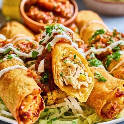 A plate of crispy chicken taquitos topped with salsa, sour cream, and chopped cilantro, served on a bed of shredded lettuce, with extra salsa and herbs in the background.
