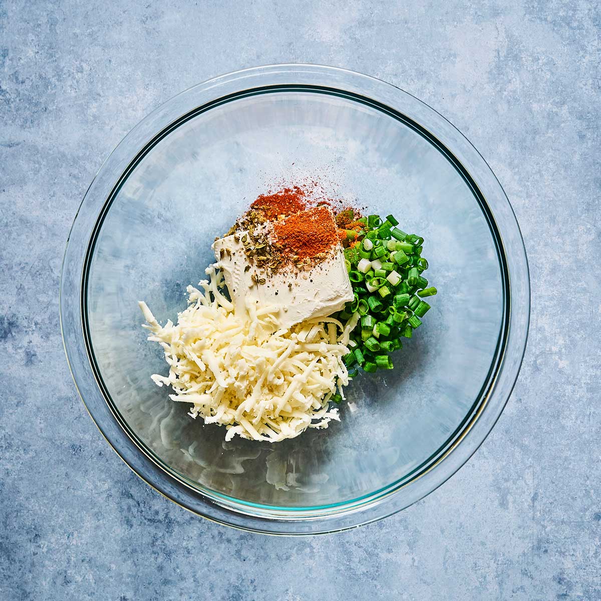 A glass bowl containing shredded cheese, a block of cream cheese, chopped green onions, and assorted spices on a light blue surface.