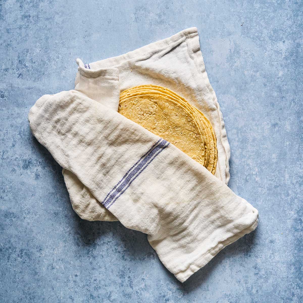 A stack of corn tortillas partially wrapped in a white cloth with a blue stripe, placed on a light blue textured surface.
