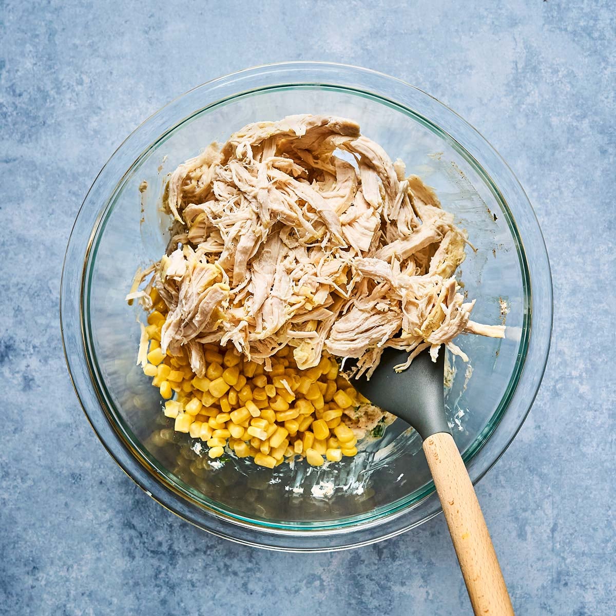 A glass bowl contains shredded chicken and yellow corn, with a gray spatula resting inside. The bowl is placed on a light blue textured surface.