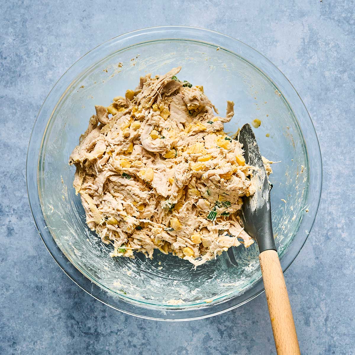 A glass bowl filled with a mixture of shredded chicken, chickpeas, and herbs. A spatula with a wooden handle rests in the bowl, all set on a light blue textured surface.