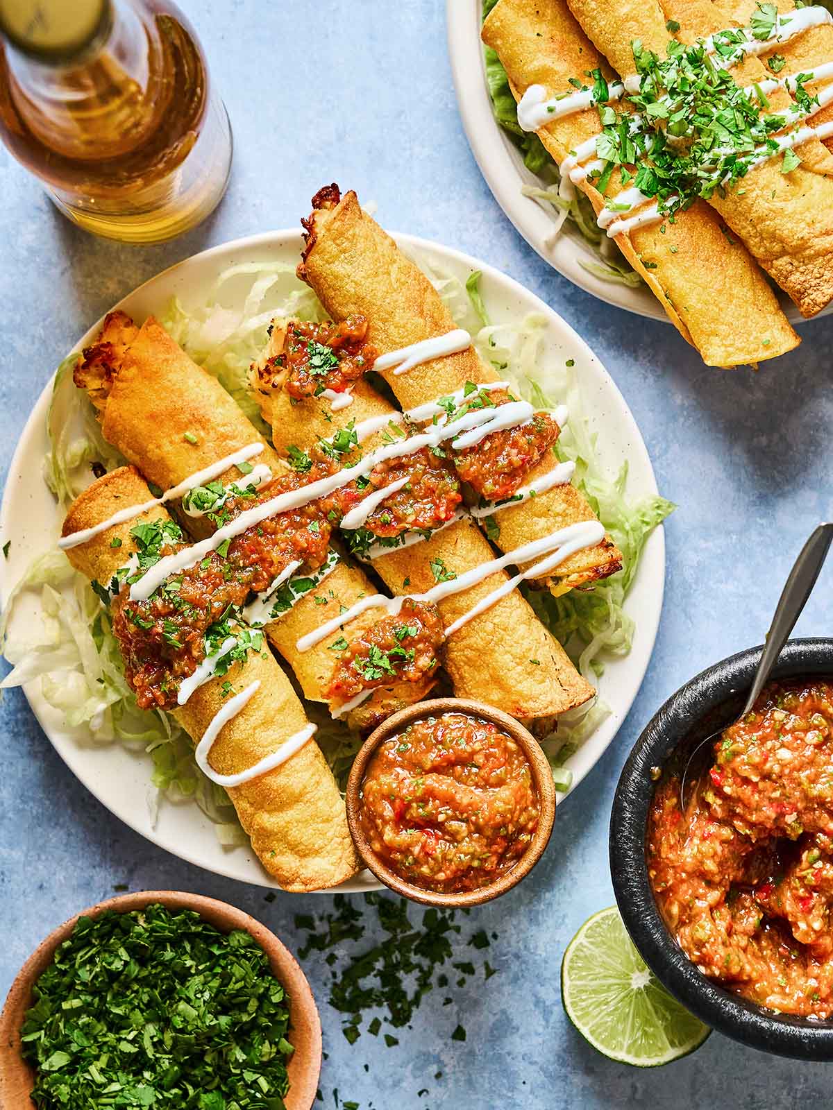 A plate of crispy taquitos topped with salsa, sour cream, and chopped cilantro, served on shredded lettuce with salsa in a small bowl. A beer bottle, extra taquitos, salsa, lime, and chopped cilantro are nearby.
