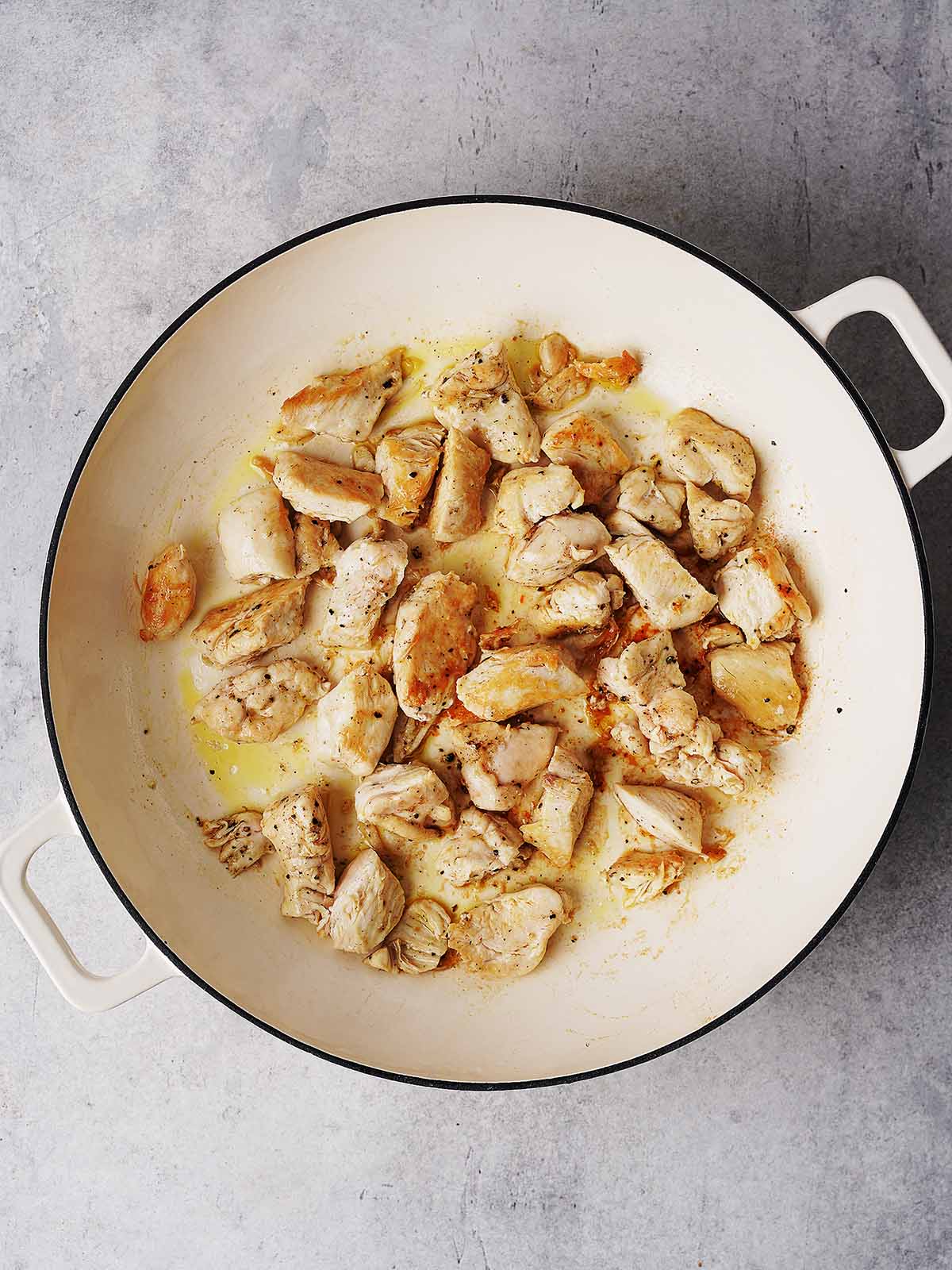 Chunks of browned chicken cooking in a large white, round pan with two handles, set on a light gray countertop.