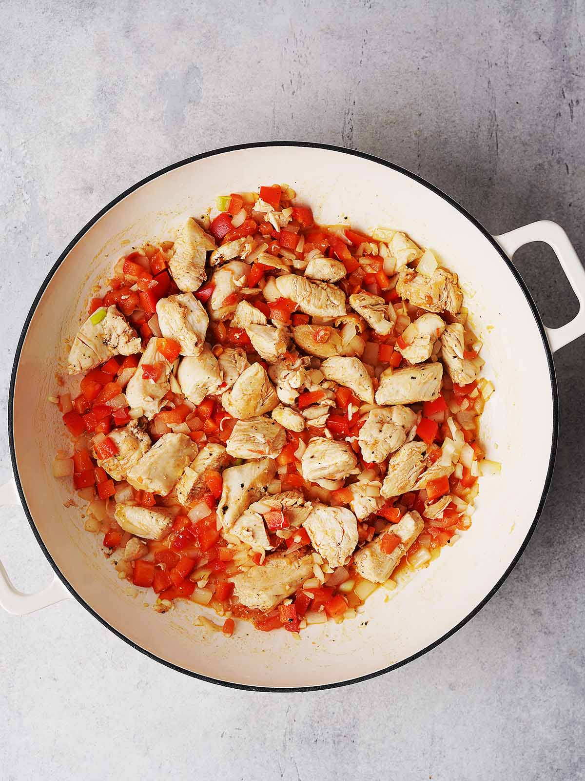 A white pot filled with cooked chicken pieces, diced red bell peppers, and chopped onions, all sautéed and mixed together on a light gray surface.