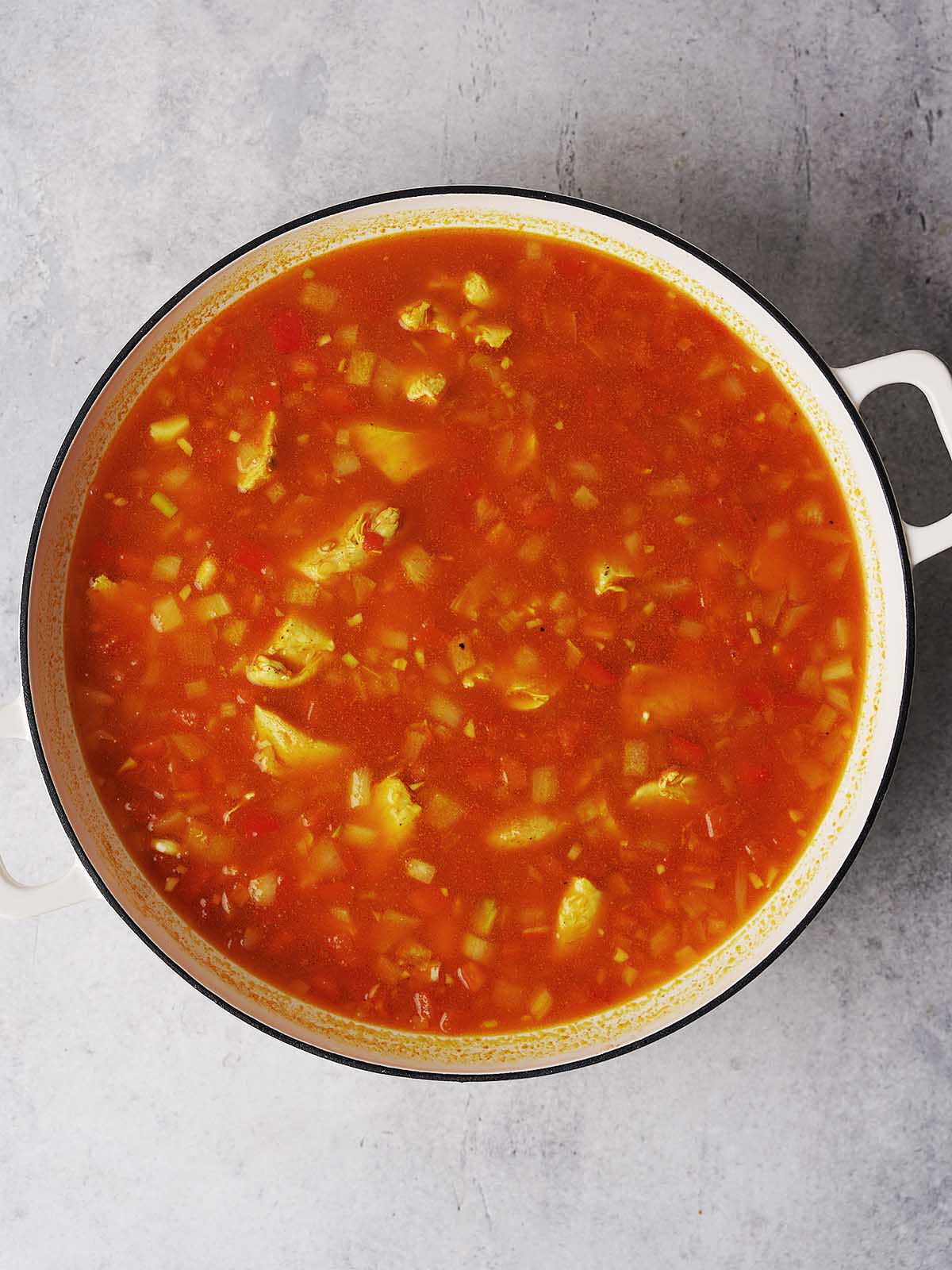 A large white pot filled with tomato-based vegetable soup, containing visible chunks of vegetables, sits on a light gray surface.
