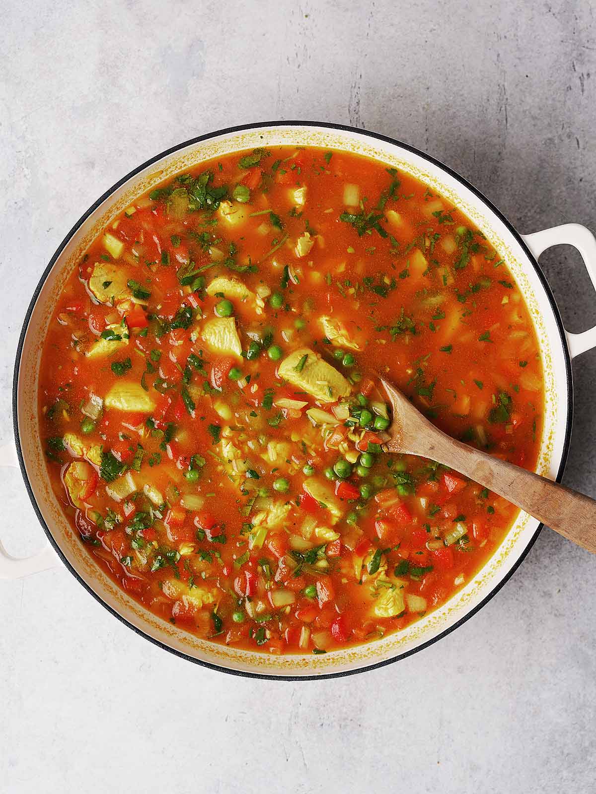 A pot of vegetable soup with visible chunks of avocado, tomatoes, green peas, potatoes, and herbs, being stirred with a wooden spoon on a light-colored surface.