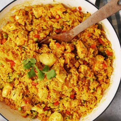A white pot filled with yellow rice, chicken pieces, peas, and carrots. A wooden spoon rests inside the pot, with cilantro garnishing the rice. A blue checkered cloth is nearby.