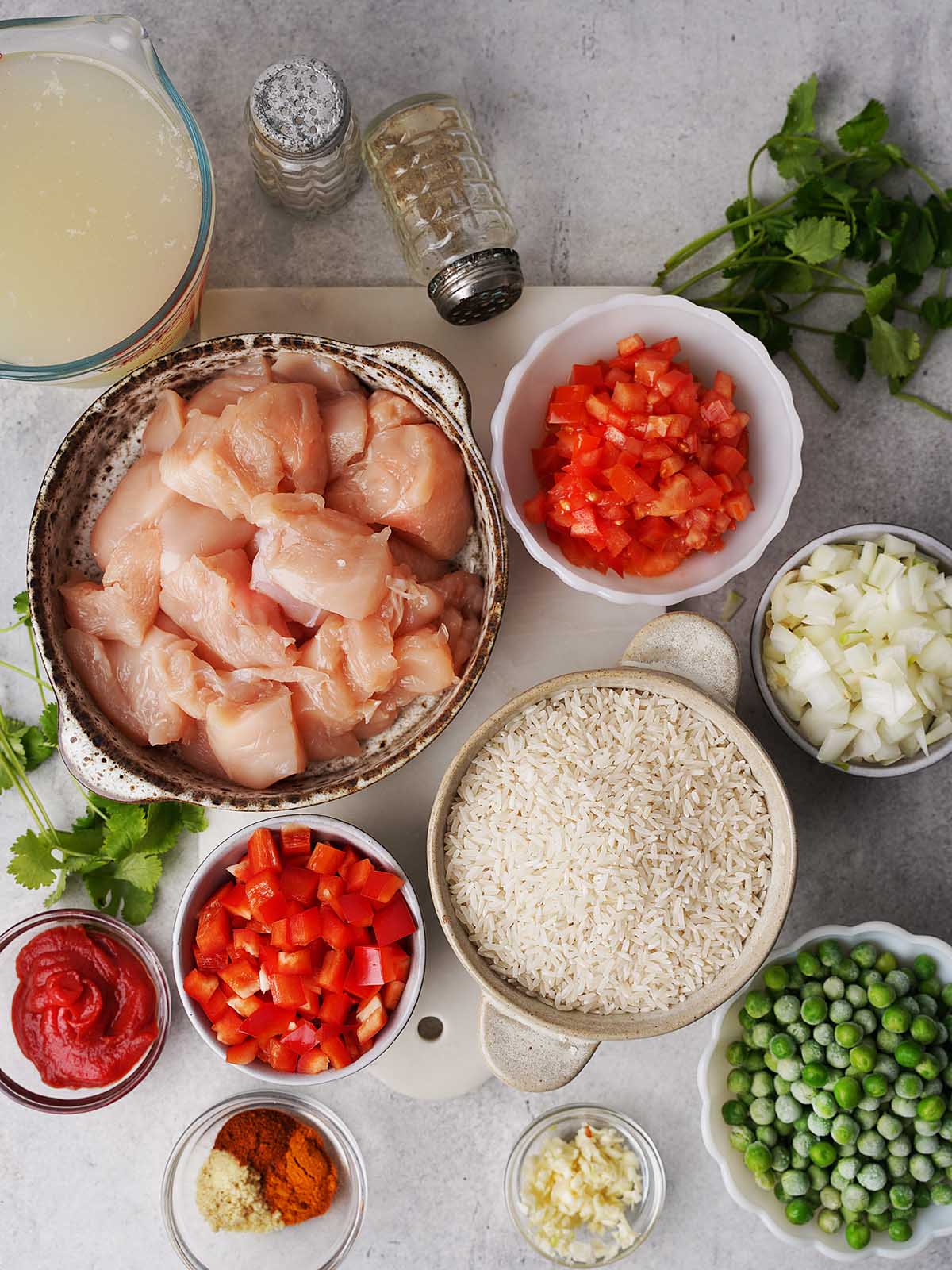 A top-down view of raw diced chicken in a bowl surrounded by ingredients in separate bowls: uncooked rice, diced tomatoes, chopped onions, red bell peppers, green peas, tomato paste, minced garlic, spices, chicken broth, salt, and fresh cilantro.