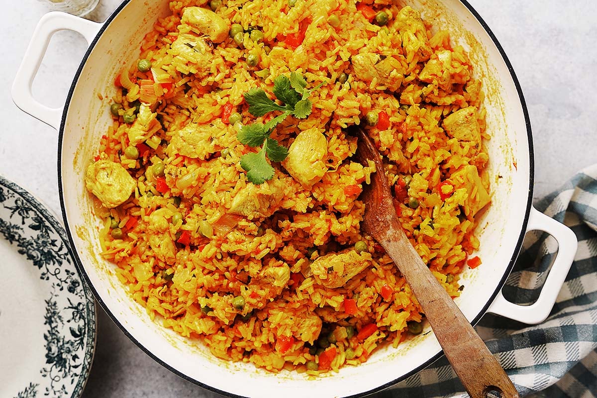 A large white pot filled with yellow rice, chunks of chicken, peas, and diced carrots, garnished with cilantro. A wooden spoon rests inside the pot, and a checkered cloth is nearby.