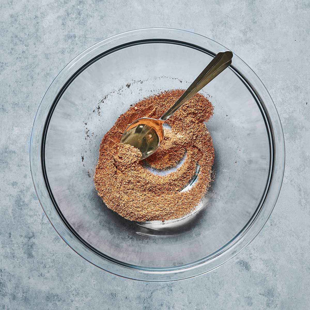 A clear glass bowl containing ground flaxseed with a gold-colored spoon resting inside, placed on a light gray textured surface.