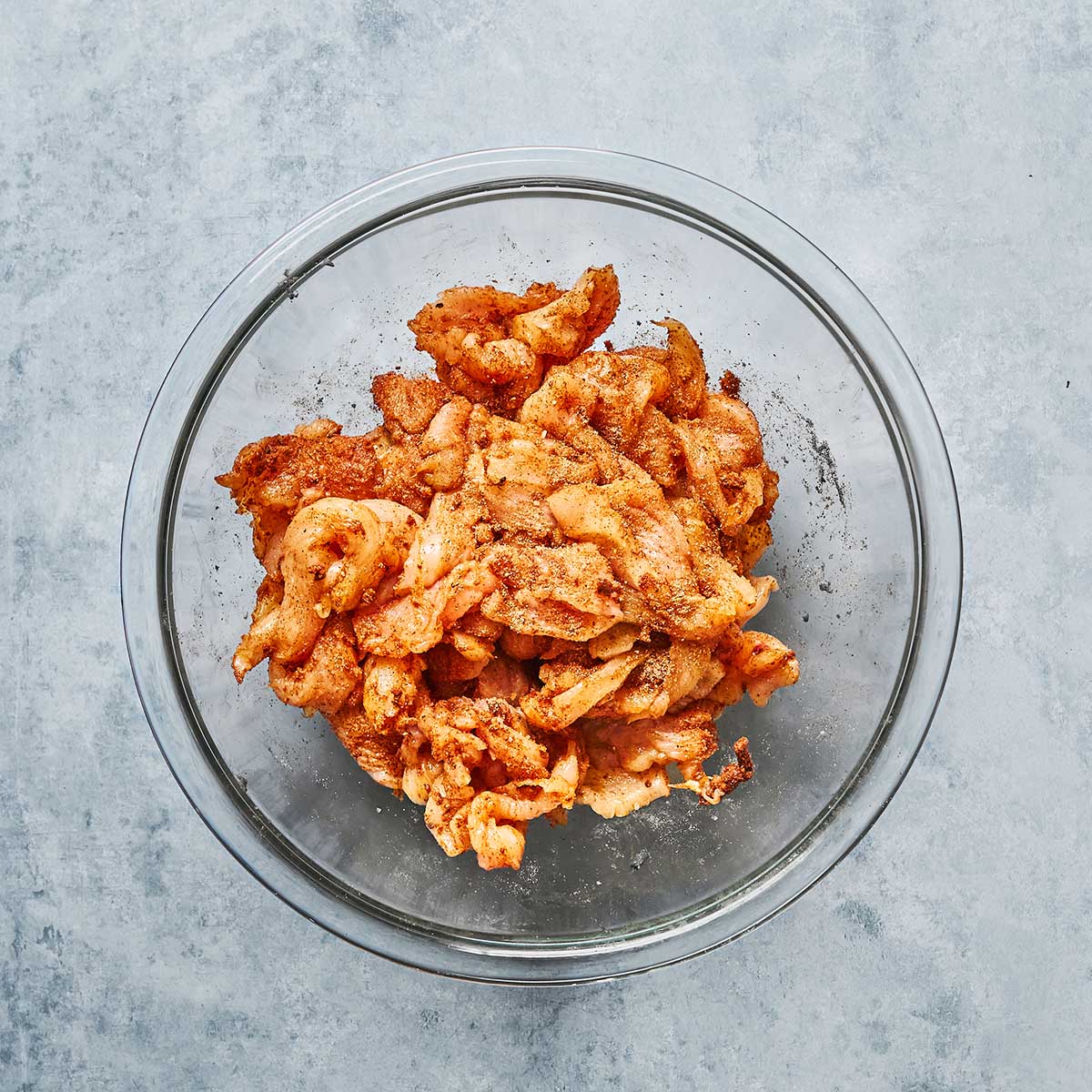 Raw, sliced chicken pieces coated with spices are in a clear glass bowl, sitting on a light gray textured surface.