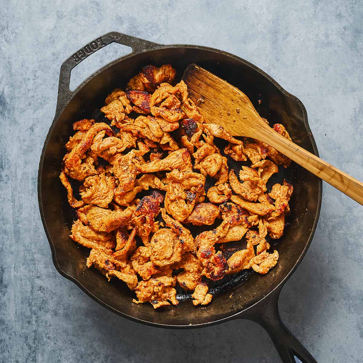 Cooked, seasoned chicken pieces in a cast iron skillet, being stirred with a wooden spatula on a gray surface.