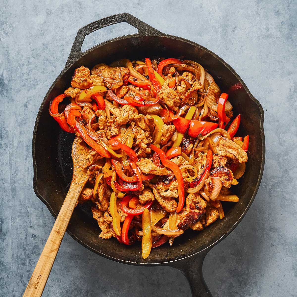 A cast iron skillet filled with cooked strips of chicken, red bell peppers, and onions. A wooden spoon rests inside the pan, mixing the colorful ingredients against a gray background.