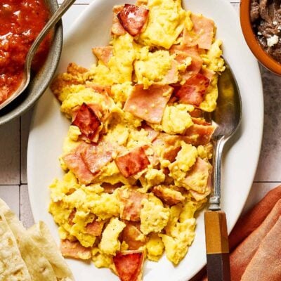 A white plate with scrambled eggs and pieces of ham, served with a spoon. Surrounding the plate are bowls of salsa, refried beans, tortillas, and a cloth napkin.