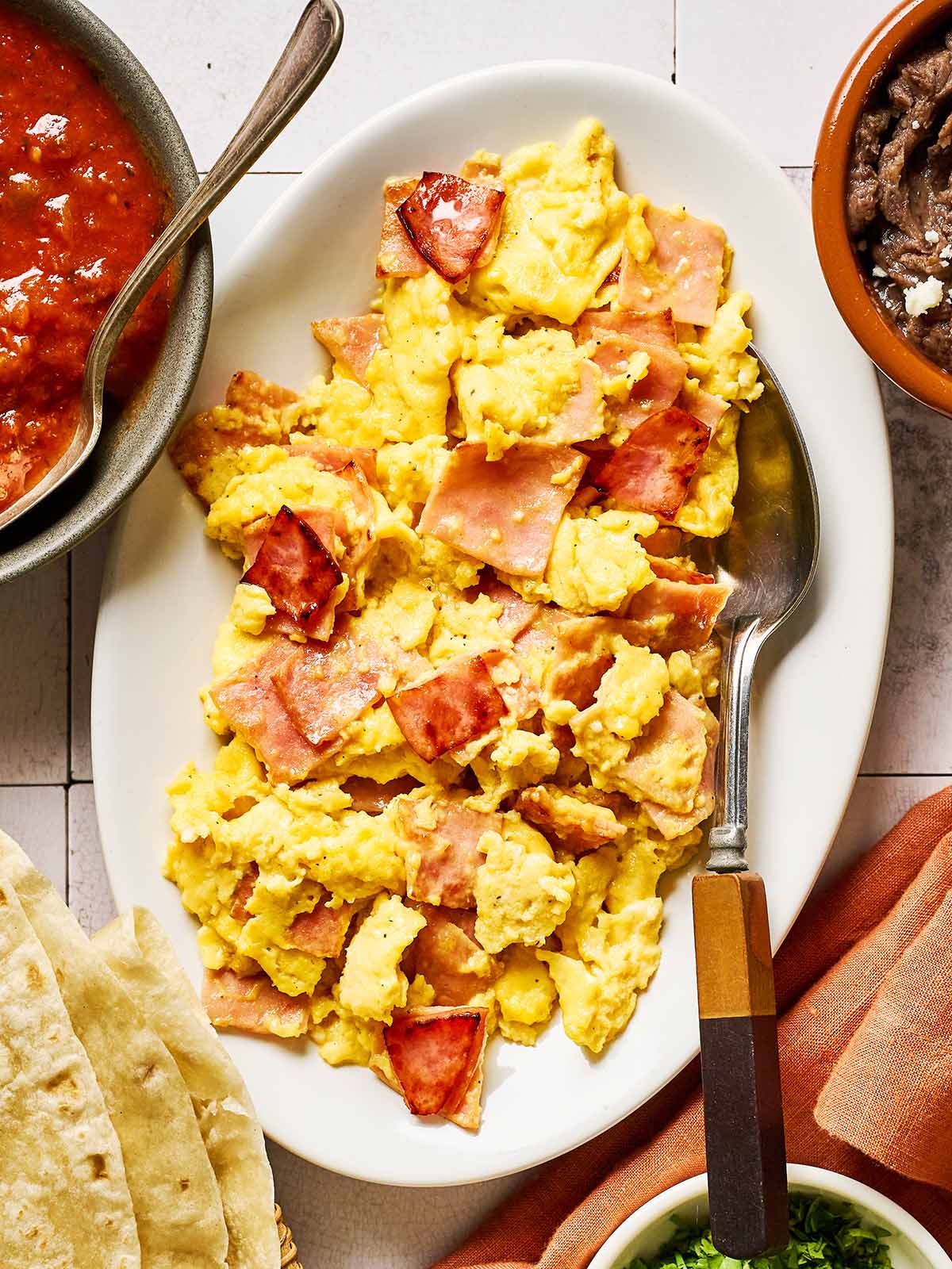 A white plate with scrambled eggs and pieces of ham, served with a spoon. Surrounding the plate are bowls of salsa, refried beans, tortillas, and a cloth napkin.