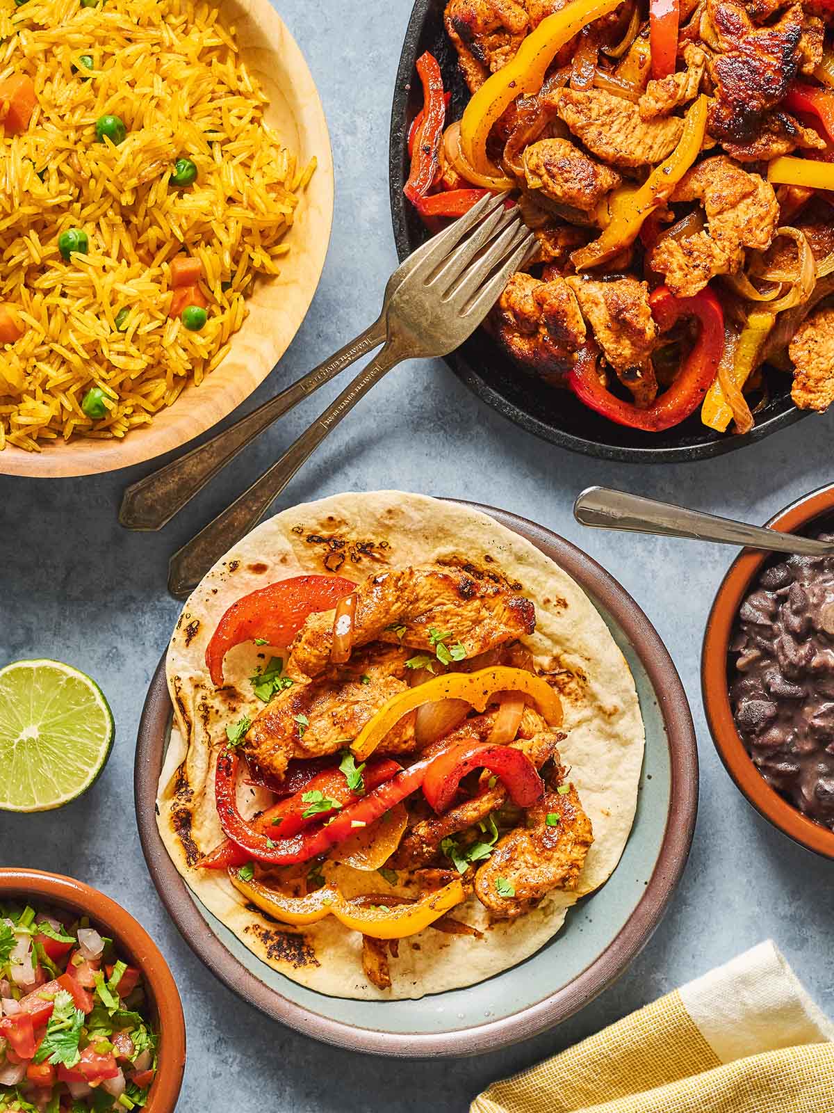 A plate with sauteed chicken on a tortilla, topped with sautéed bell peppers and herbs, surrounded by bowls of yellow rice, black beans, pico de gallo, and a lime half. A fork and knife rest nearby.