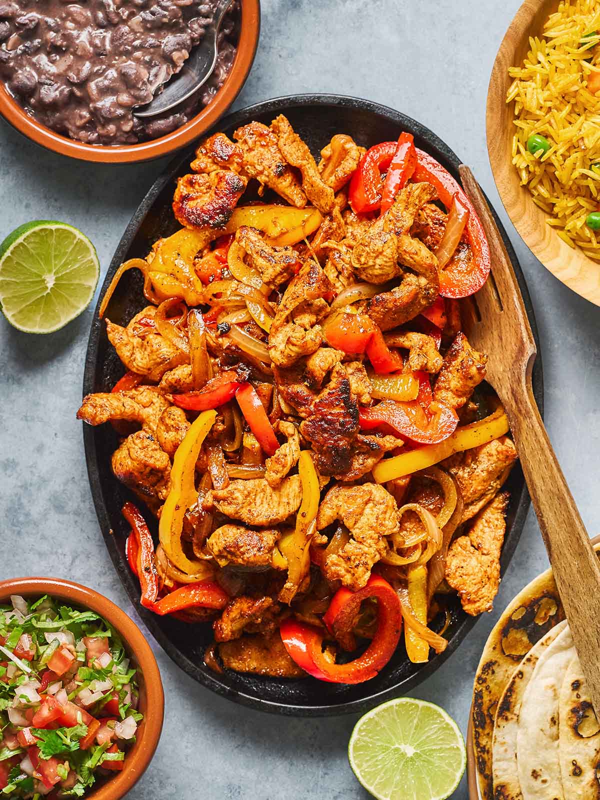 A sizzling skillet of fajitas with grilled chicken strips, red and yellow bell peppers, and onions, surrounded by sides of rice, black beans, pico de gallo, lime halves, and tortillas.