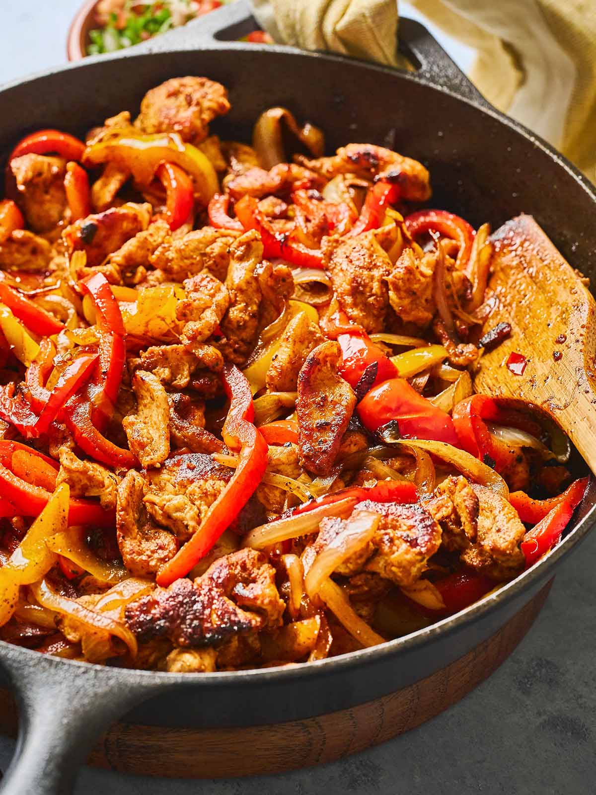 A sizzling skillet filled with cooked chicken strips, red and yellow bell peppers, and onions, all seasoned and sautéed, with a wooden spatula resting in the pan.