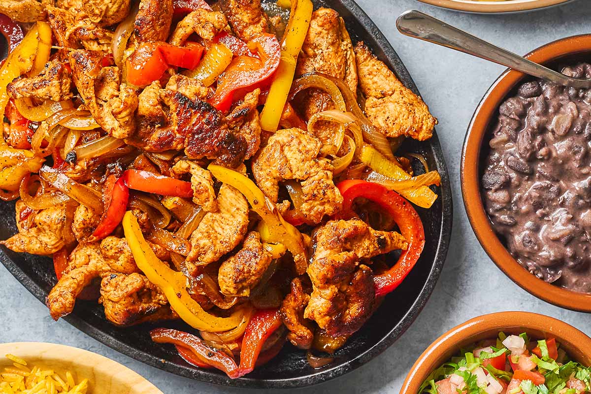 A sizzling skillet of chicken fajitas with grilled chicken strips, red and yellow bell peppers, and onions is served with sides of black beans and pico de gallo on a gray tabletop.