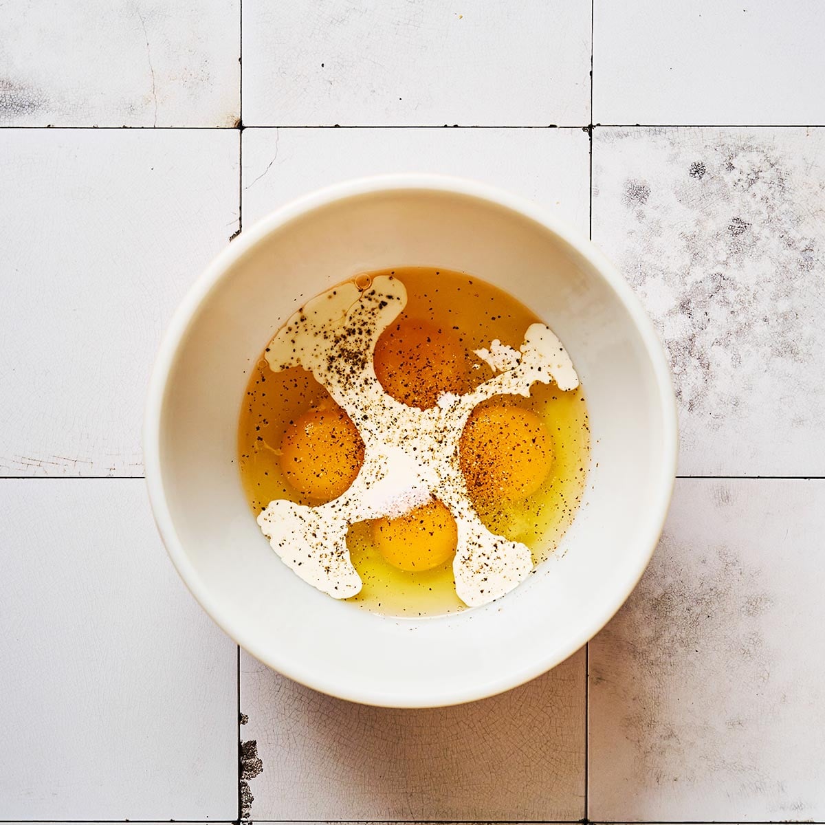 A white bowl containing three cracked eggs, some cream, and a sprinkle of black pepper sits on a worn white tiled surface.