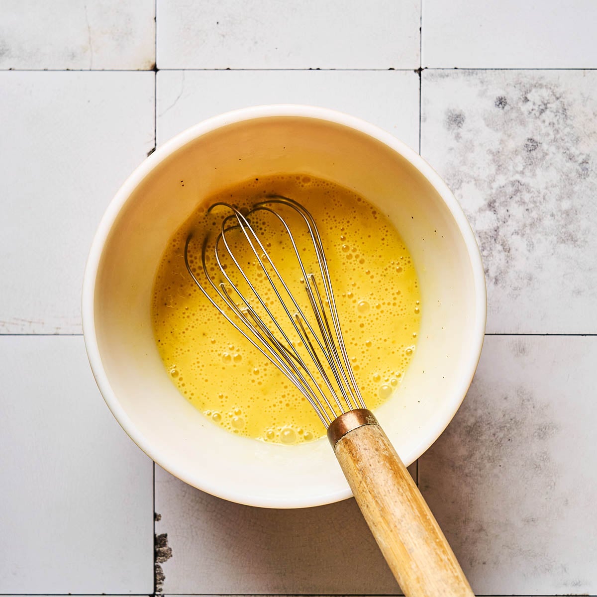 A metal whisk with a wooden handle rests in a white bowl containing beaten eggs, placed on a white tiled surface with some stains and discoloration.