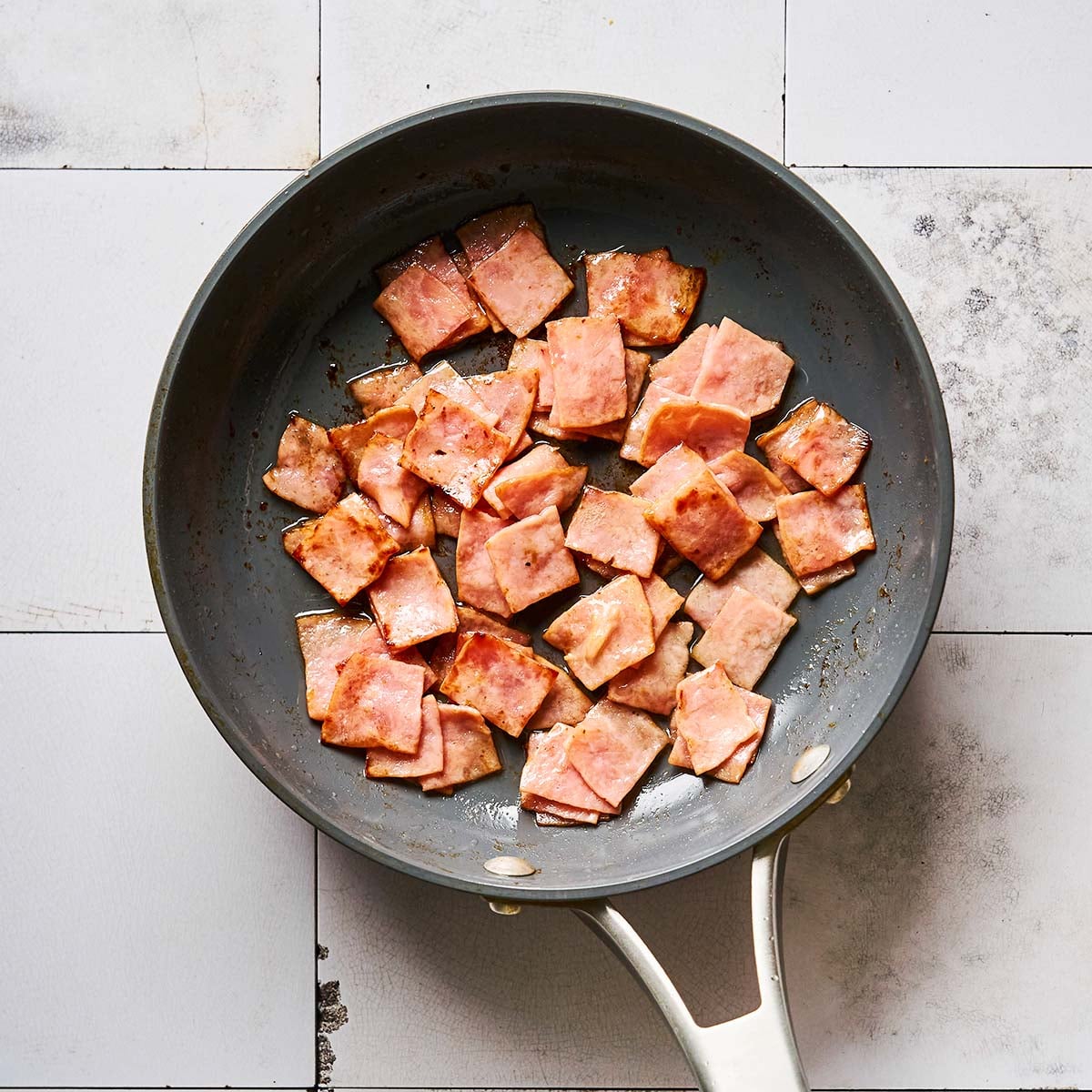 A nonstick frying pan with diced pieces of cooked ham sits on a white tiled surface.