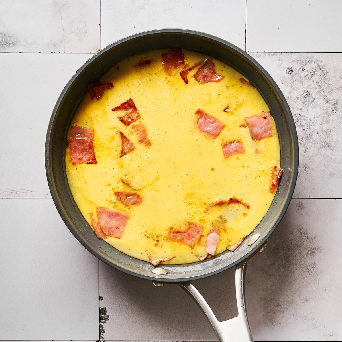 A frying pan on a tiled surface contains an omelette with visible pieces of cooked ham. The omelette is mostly set, with a smooth yellow surface and scattered pink ham pieces.