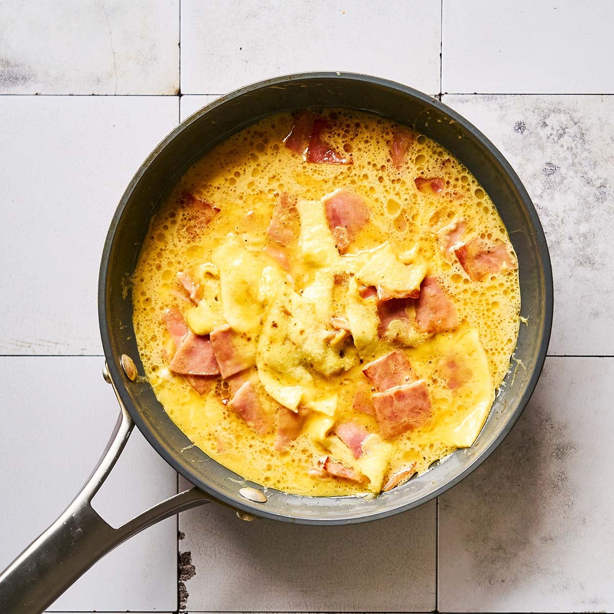 A frying pan on a tiled surface, filled with scrambled eggs mixed with chunks of ham and partially melted cheese.