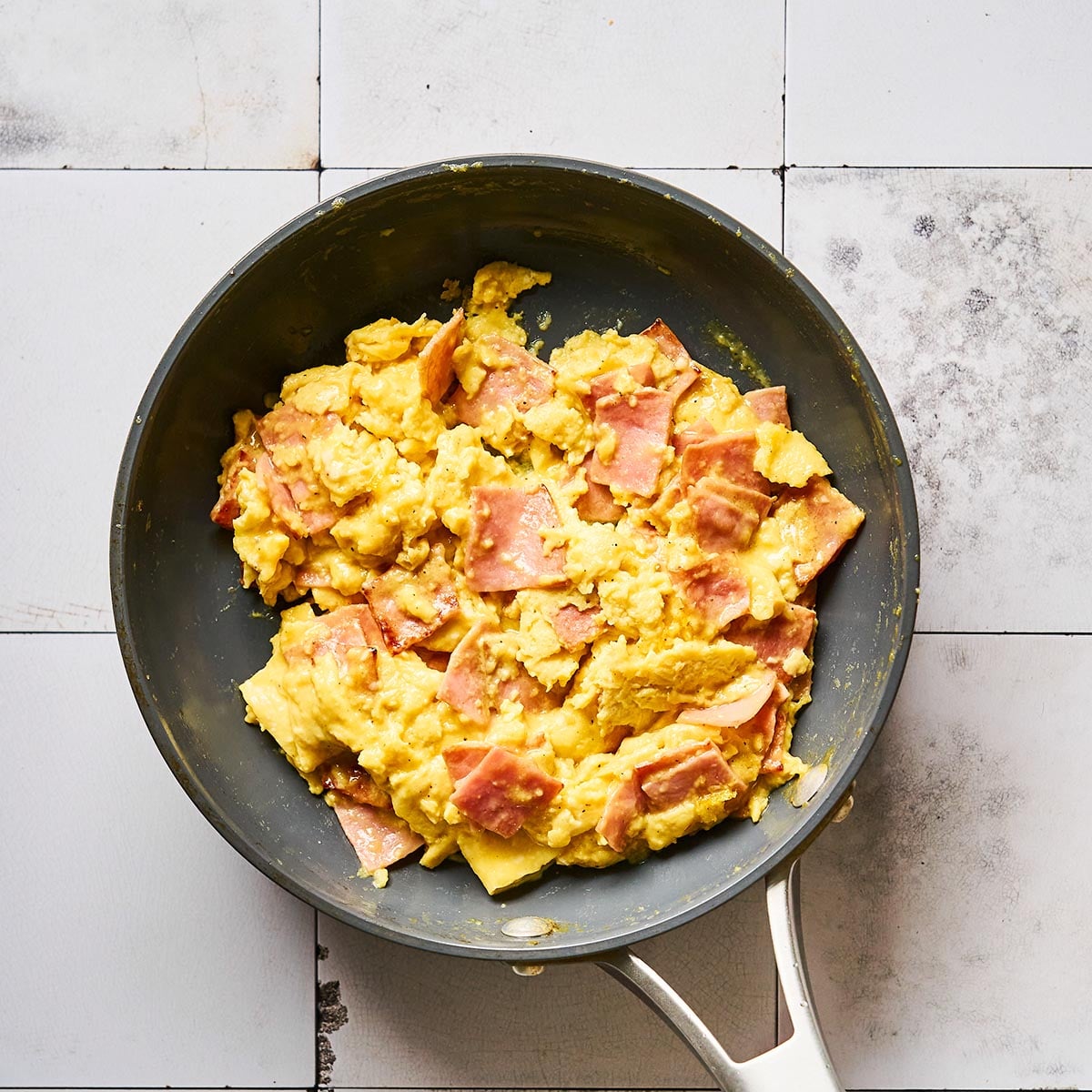 A frying pan on a white tiled surface contains scrambled eggs mixed with pieces of ham.