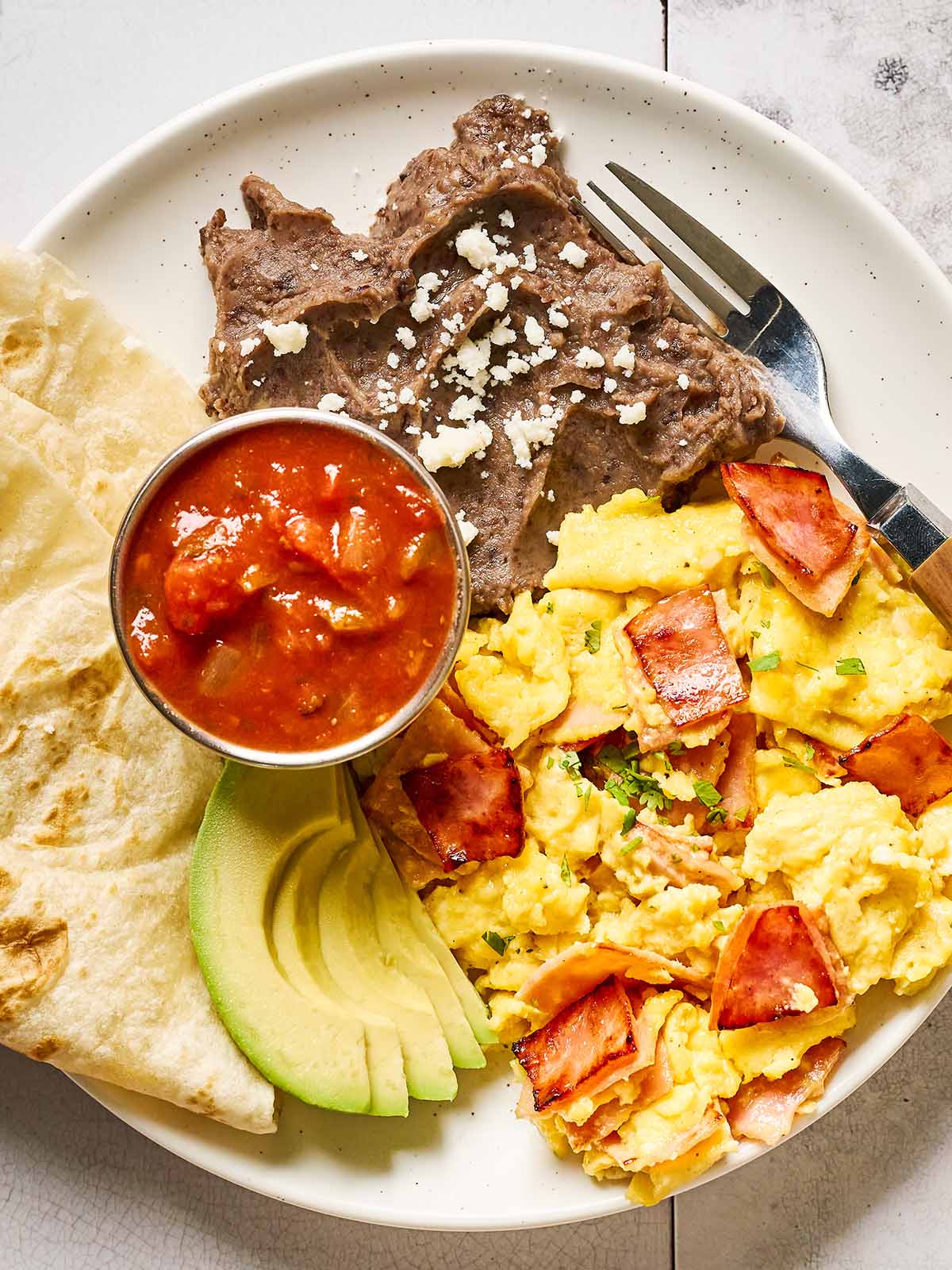 A plate with scrambled eggs and pieces of ham, grilled steak topped with cheese, refried beans, sliced avocado, tortillas, and a small cup of salsa. A fork rests on the plate.