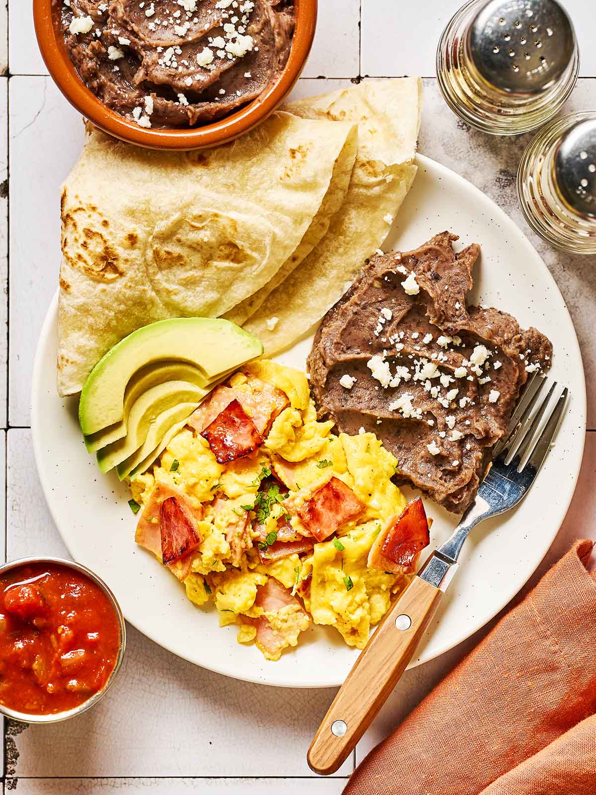 A plate with scrambled eggs mixed with tomatoes and ham, sliced avocado, refried beans topped with cheese, and folded flour tortillas. Salsa is in a small bowl beside the plate, along with salt and pepper shakers.