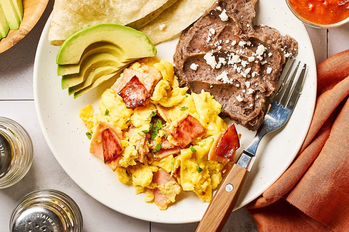 A plate with scrambled eggs and ham, grilled steak topped with cheese, sliced avocado, tortillas, and a fork. There’s an orange napkin, salsa, and salt and pepper shakers nearby.