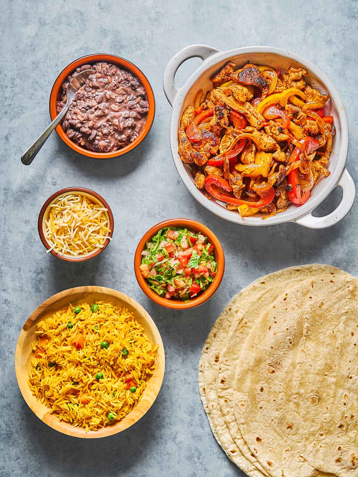 Overhead view of a spread with flour tortillas, a dish of sautéed peppers and chicken, bowls of yellow rice, black beans, shredded cheese, and fresh salsa on a gray surface.