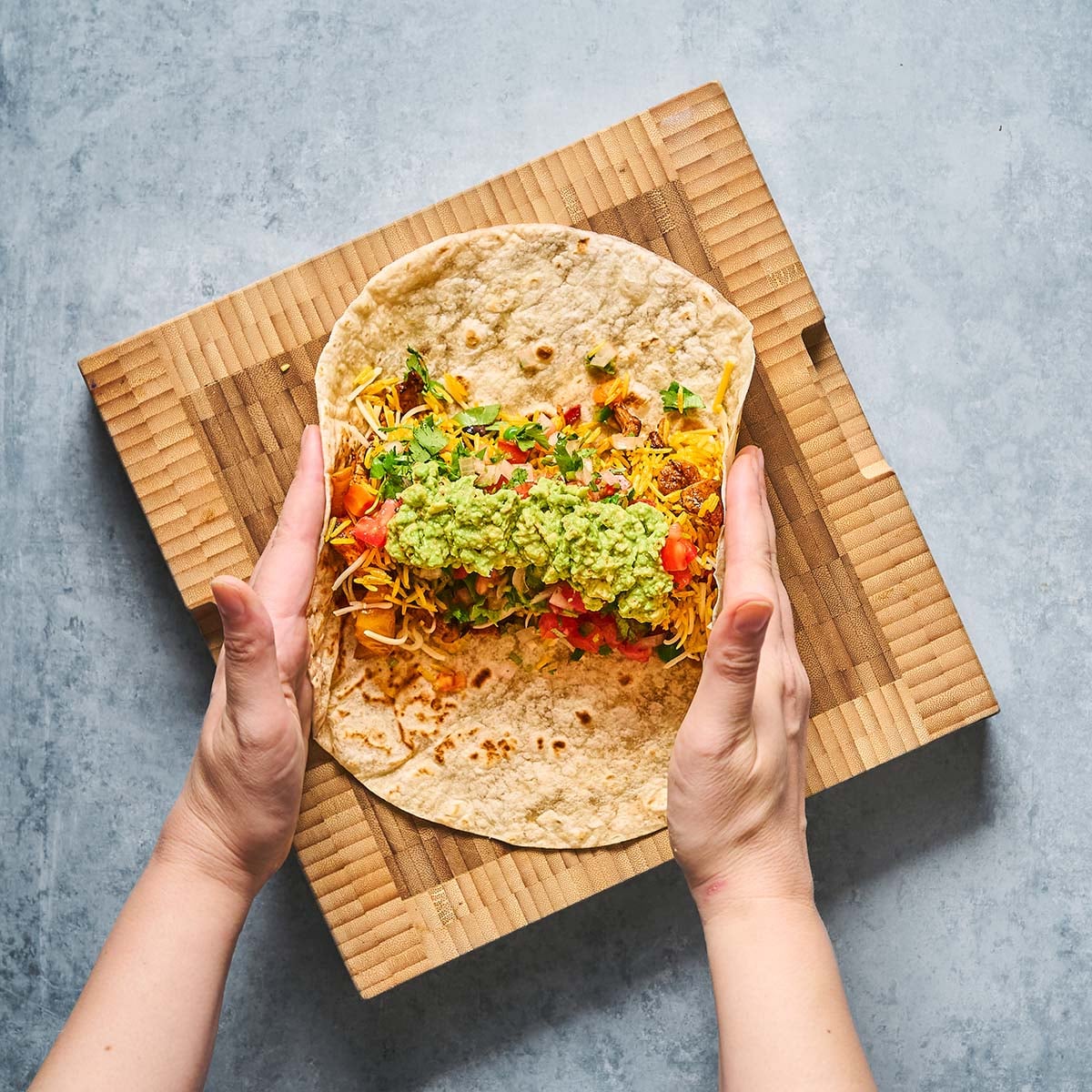 Two hands are folding a tortilla filled with shredded cheese, chopped tomatoes, greens, and guacamole on a wooden cutting board.