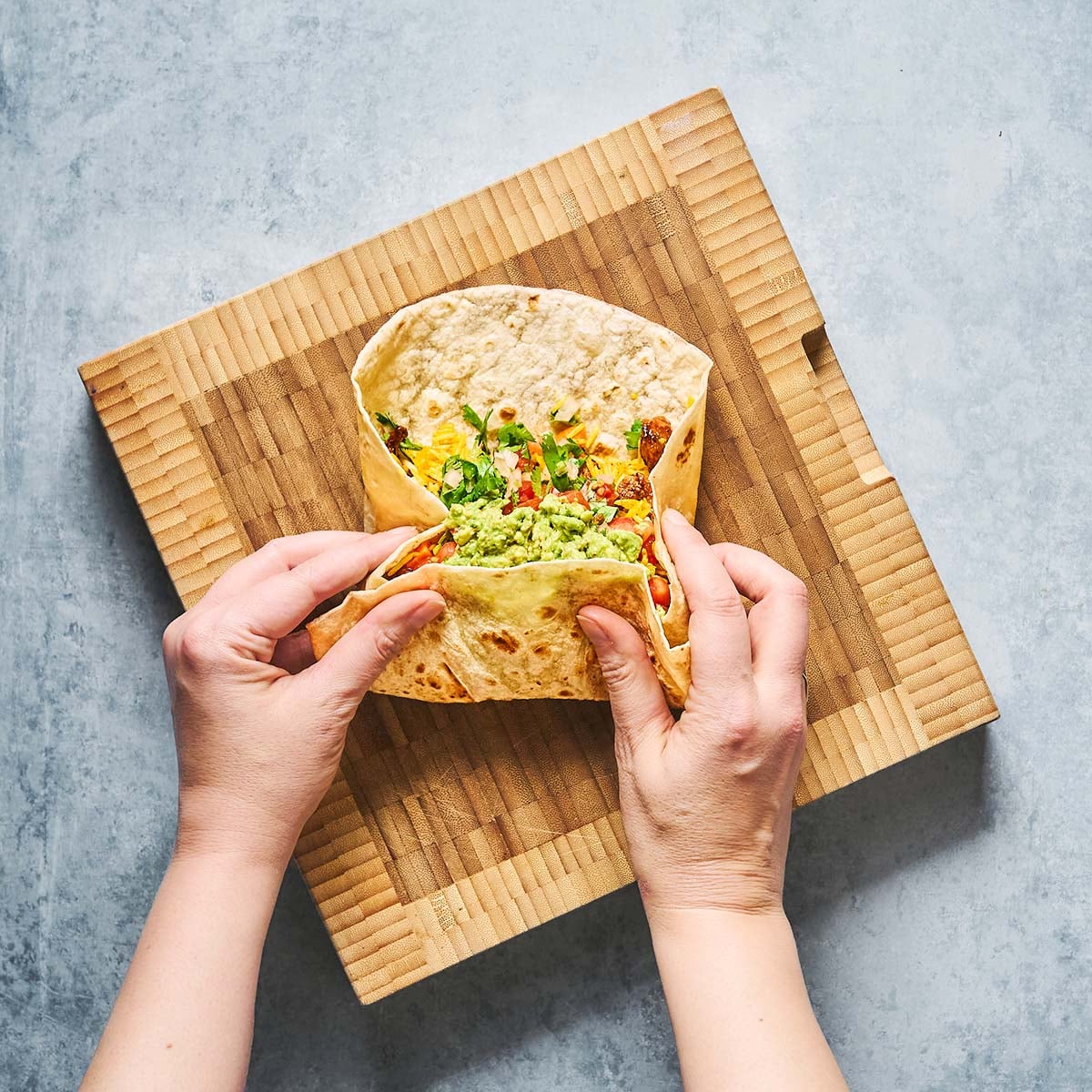 Two hands folding a tortilla wrap filled with guacamole and vegetables on a wooden cutting board, viewed from above.