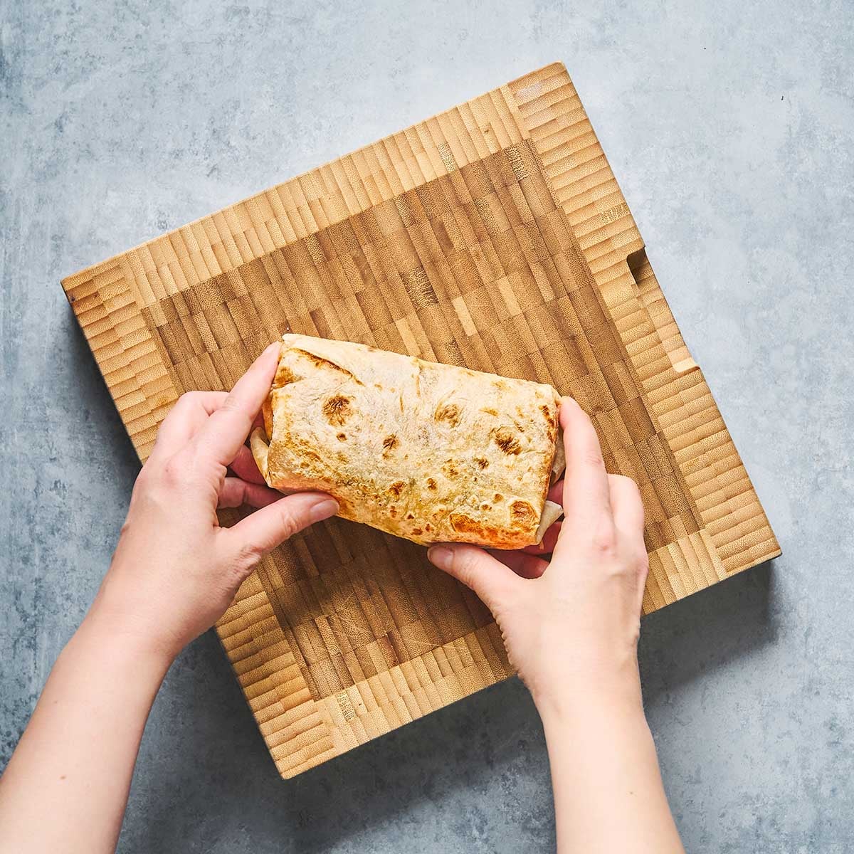 Two hands hold a wrapped sandwich or burrito above a rectangular wooden cutting board on a gray surface.
