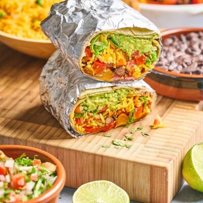 Two burritos wrapped in foil, cut in half to show fillings of guacamole, lettuce, tomato, cheese, and beans, stacked on a wooden board with lime, salsa, black beans, and rice in the background.