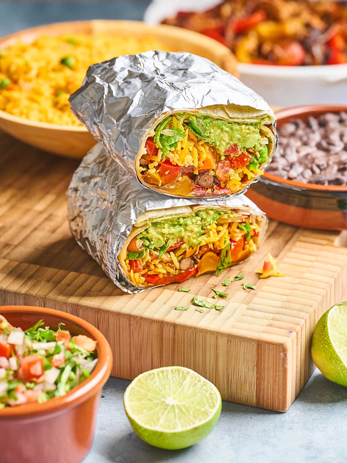 Two burritos wrapped in foil, cut in half to show fillings of guacamole, lettuce, tomato, cheese, and beans, stacked on a wooden board with lime, salsa, black beans, and rice in the background.