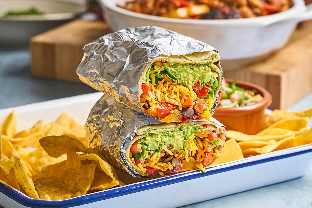 A foil-wrapped burrito cut in half, filled with rice, beans, vegetables, and guacamole, sits on a tray with tortilla chips. A small bowl of salsa and a dish of mixed food are in the background.