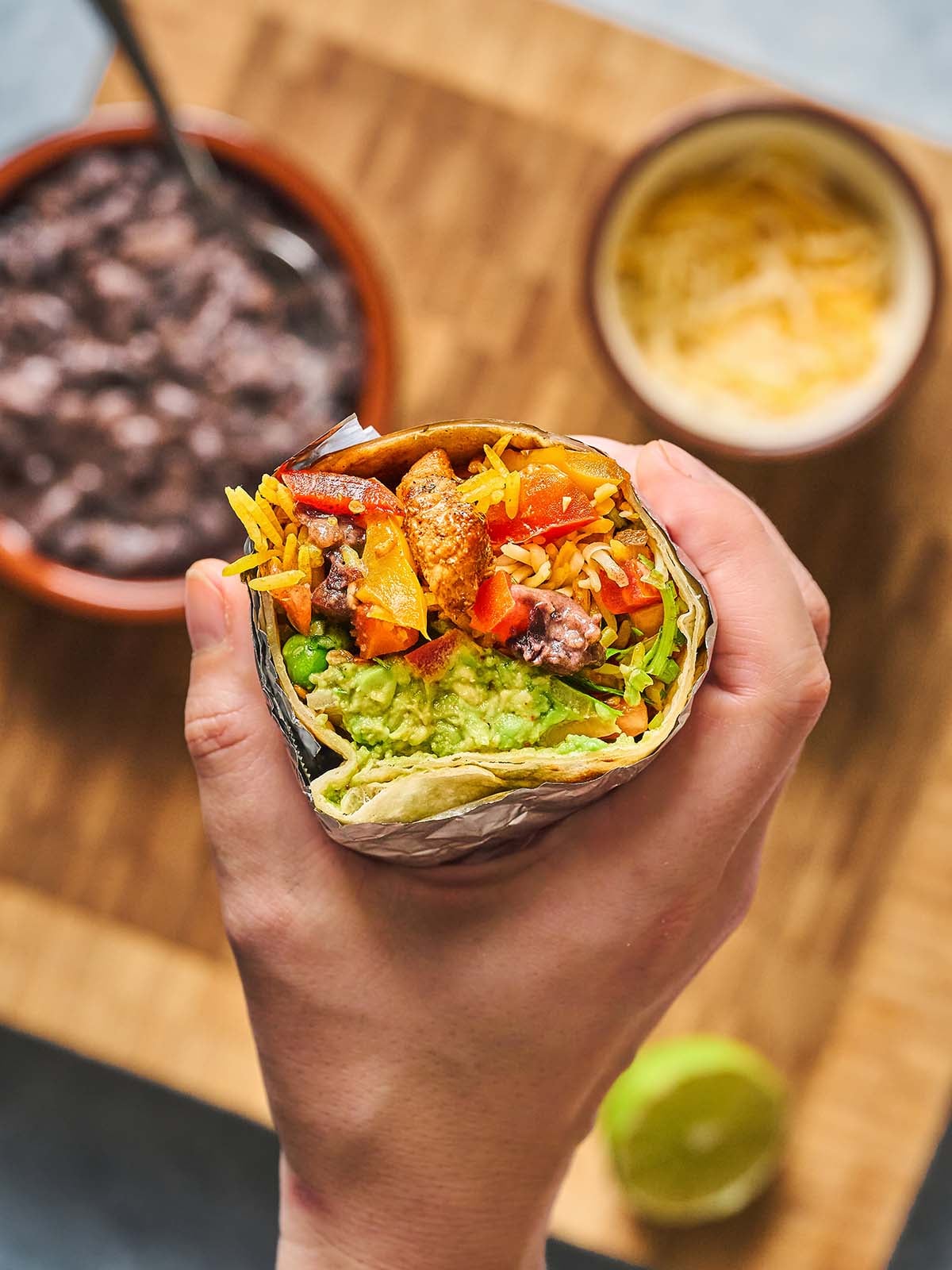 A hand holds a burrito filled with guacamole, rice, beans, chicken, cheese, and sautéed peppers. In the background, bowls of black beans and shredded cheese sit on a wooden board with a lime nearby.