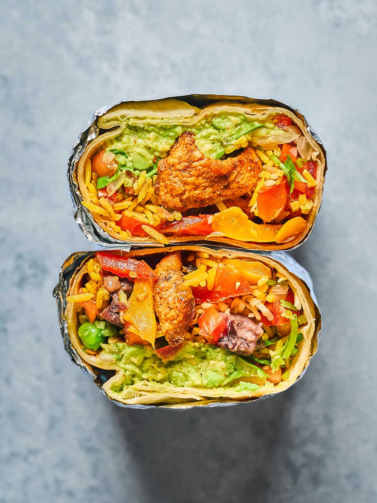 A close-up of two halves of a burrito wrapped in foil, filled with rice, vegetables, guacamole, and pieces of cooked chicken, displayed on a gray surface.