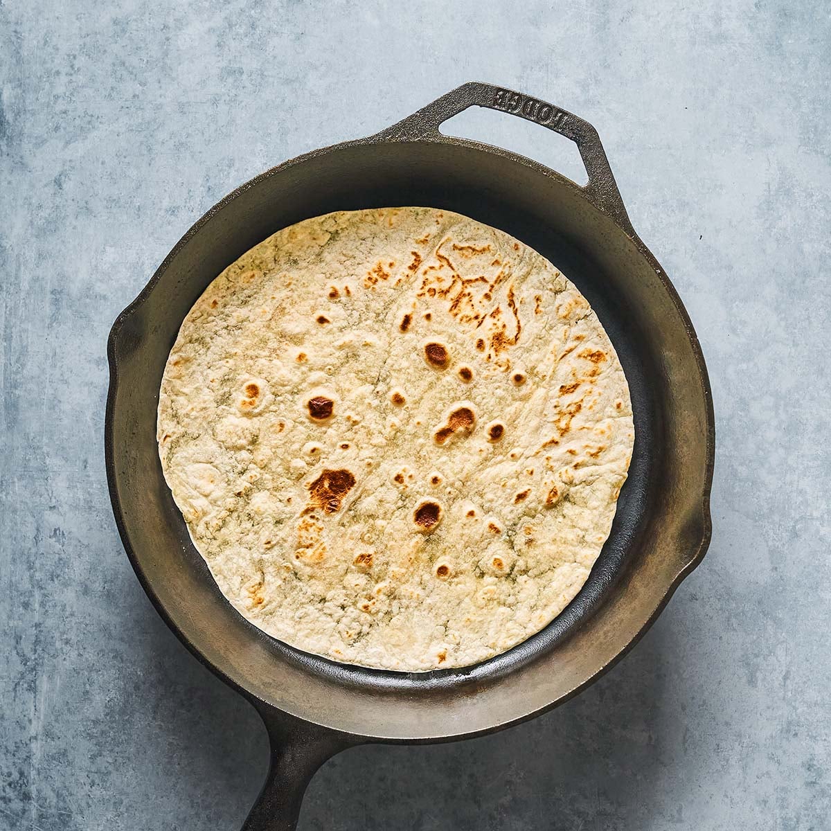 A round, lightly browned flatbread cooking in a black cast iron skillet, set on a textured gray surface.