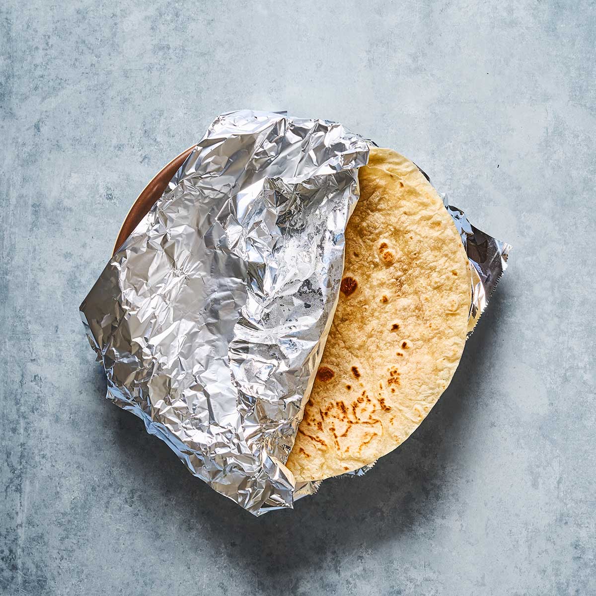 A round tortilla partially wrapped in crumpled aluminum foil sits on a light gray textured surface.