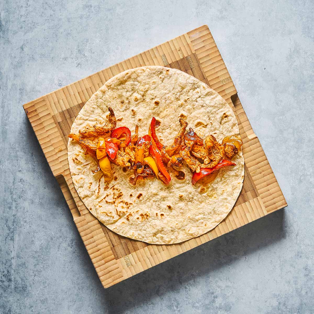 A flour tortilla topped with cooked sliced red and yellow bell peppers and onions, placed on a brown, ridged cardboard square over a gray surface.