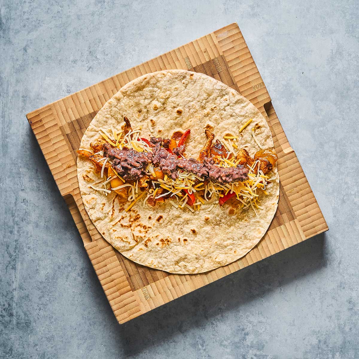A flour tortilla topped with shredded cheese, beans, and cooked vegetables sits on a piece of cardboard against a gray background.