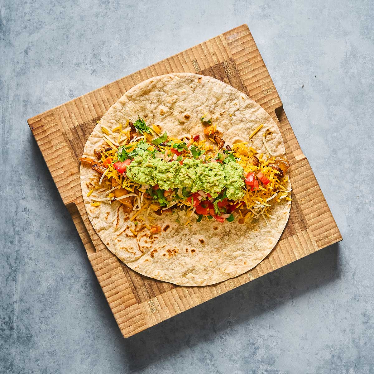 A flour tortilla filled with shredded cheese, lettuce, tomatoes, and a generous scoop of guacamole, placed on a brown textured board against a gray background.